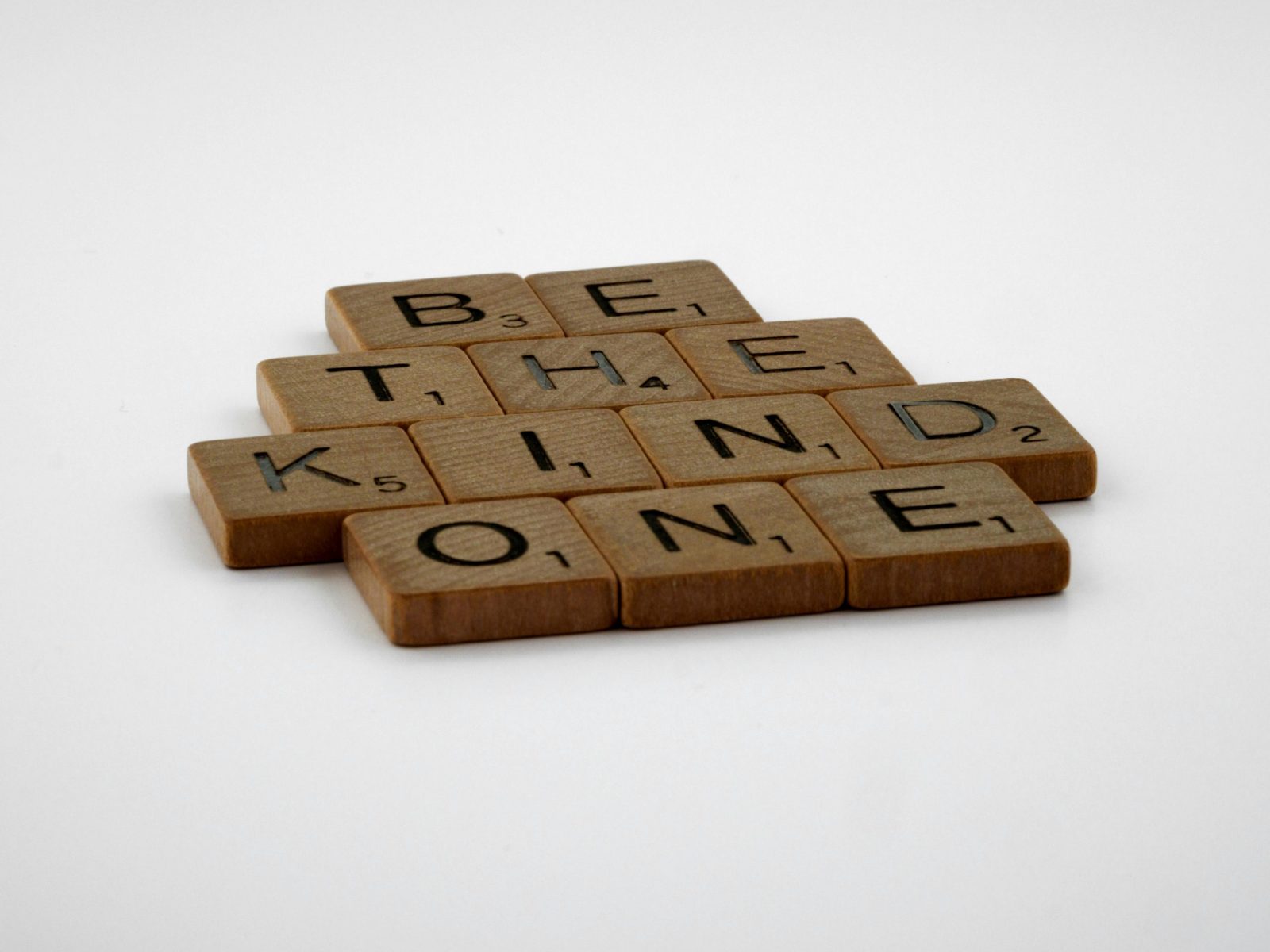 Wooden Scrabble tiles spelling 'Be The Kind One' with focus on words and white background.
